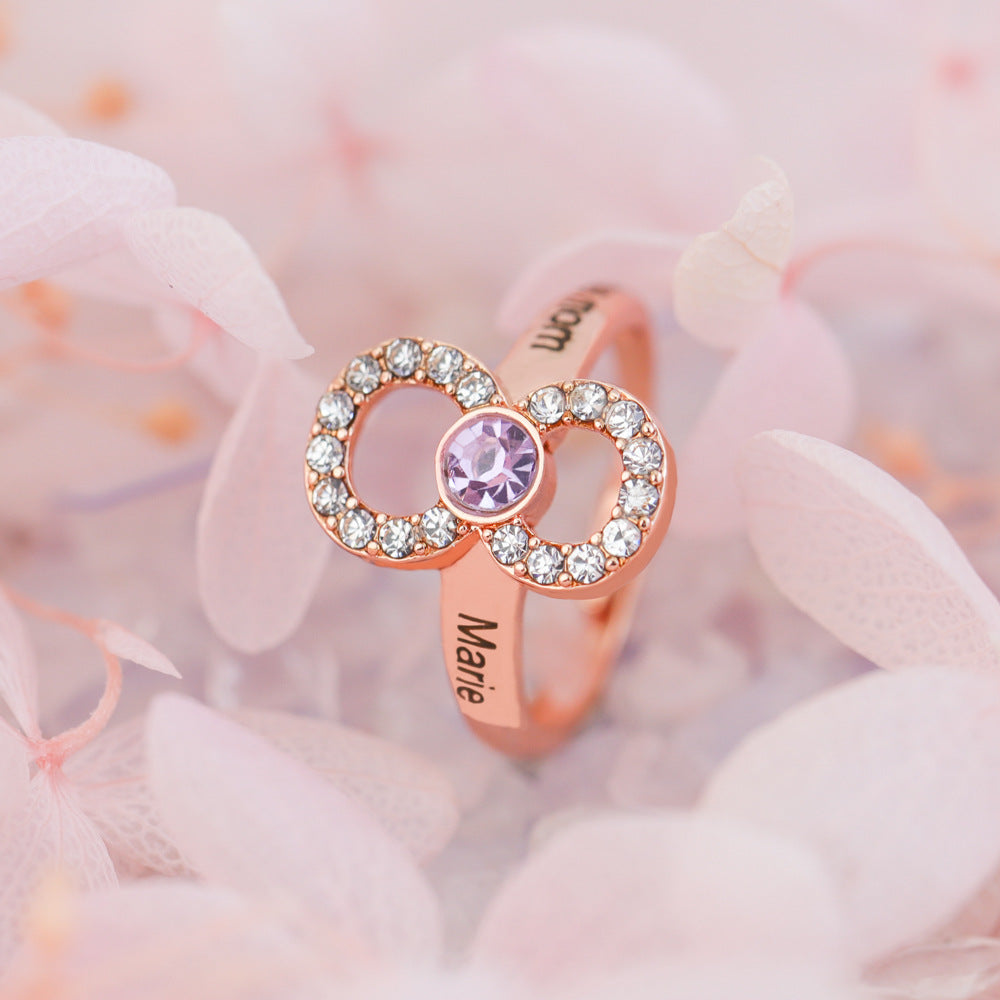 A rose gold engagement ring with a central purple diamond surrounded by smaller clear crystals, positioned on a delicate flower background.