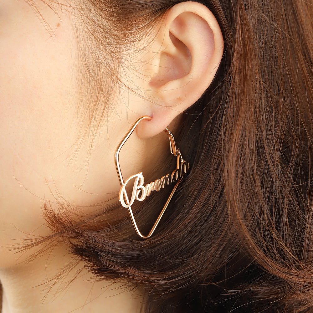 A person wearing a pair of custom-made earrings with the English name 'Brenda' inscribed in a square shape, with a rose gold plating.